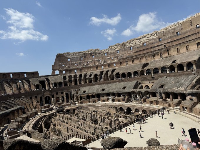 A view from inside the Roman Colosseum showing the ancient amphitheater's tiered seating, arched walls, and the exposed hypogeum (underground chambers) where visitors are walking around the arena floor.