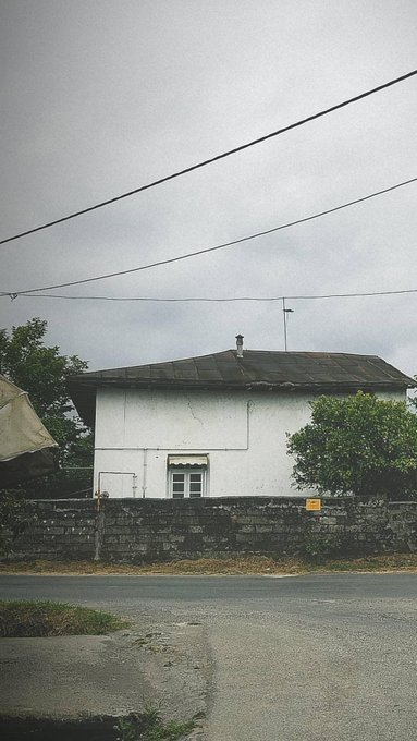 A white single-story house with a stone wall and tiled roof under an overcast sky, with a small yellow sign on the wall.