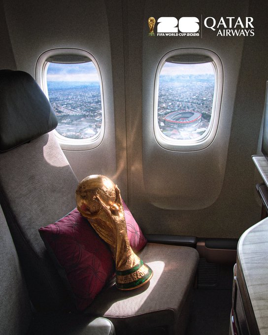 The image shows a large, gold trophy sitting on an airplane seat. The trophy appears to be a soccer ball-shaped item with a green base and gold accents. It is placed on a purple pillow that is positioned behind the seat. In the background of the photo, there are windows providing a view of a city outside the plane. The image also contains text that reads "If Sui Gets to $2 i am going to buy 2 tickets for Random person." This suggests that someone might be planning to purchase two tickets for a random person as a gift or surprise.
