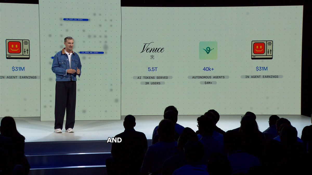 The image shows a man standing at a podium, giving a speech or presentation. The audience consists of several people sitting behind him, attentively listening to his words. The background features a large screen displaying information about a company called Venmo, which is being discussed during the event.
