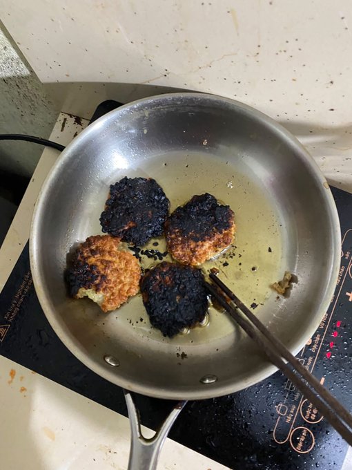 Four patties are frying in a stainless steel pan on an induction cooktop, with some pieces visibly burnt and blackened while chopsticks rest in the oil.