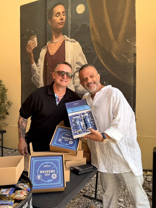 The image shows two men standing next to a table with several boxes of beer, including some Master Brews. The man on the left is holding up one of the boxes while smiling at the camera. They are both wearing sunglasses, adding a cool vibe to their appearance. The background features a painting depicting a woman and a man, which adds an artistic touch to the scene.