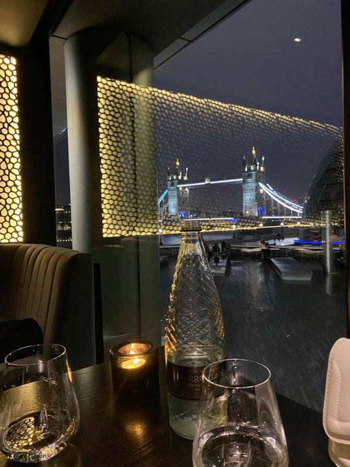 A candle-lit dinner table with wine glasses and a bottle overlooks London's illuminated Tower Bridge at night, perfectly capturing the "good night CT" sentiment with a scenic view of the Thames.