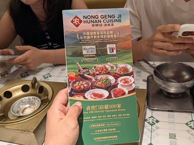 A hand holds a promotional flyer for "NONG GENG JI HUNAN CUISINE" (农耕记·湖南菜) featuring images of food, with text mentioning it is the "Leading Brand of Hunan Cuisine in China" and "Over 100 Directly Operated Stores."