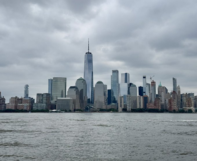 A cloudy-day photograph of the Lower Manhattan skyline featuring One World Trade Center, viewed from across the Hudson River.