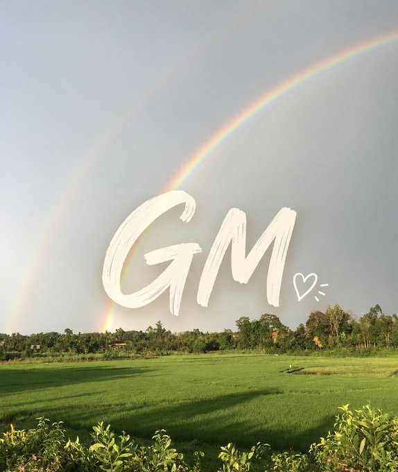 A green grassy field with trees in the background under a gray sky featuring a double rainbow, overlaid with the handwritten text 'GM' and a small heart doodle.