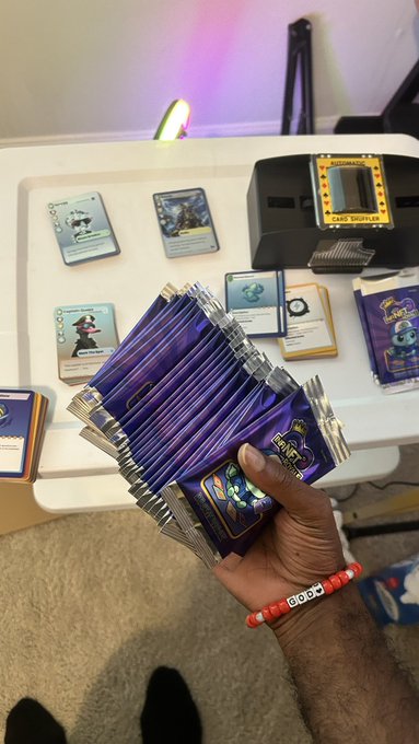 A hand holds a fan of purple trading card booster packs over a table displaying various cards, a 'CARD SHUFFLER' device, and a wristband reading 'DODO'.