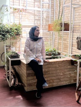 A photograph of a smiling woman wearing a hijab and white shirt, sitting on a wooden bench in what appears to be a greenhouse or plant-filled indoor space with wooden slatted walls.