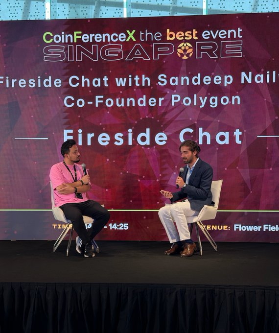 Two men sit on stage at CoinFerenceX Singapore for a 'Fireside Chat with Sandeep Nail, Co-Founder Polygon', with one in a pink shirt and the other in a dark blazer and white pants.