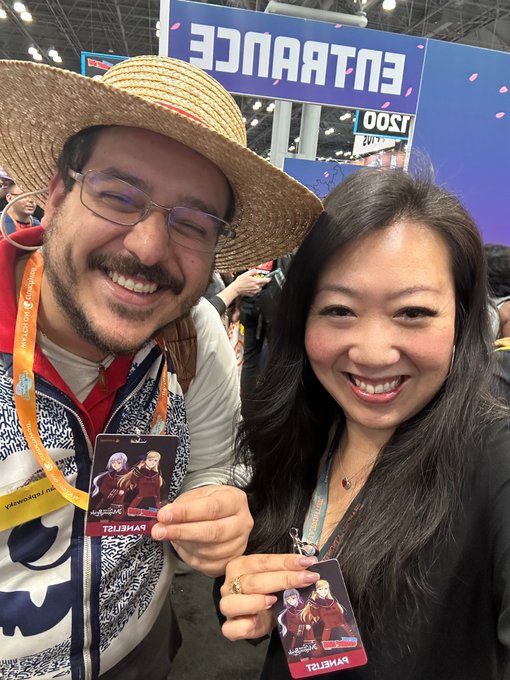 A man in a straw hat and a woman smile at the camera while holding matching character cards at what appears to be a convention, with an 'ENTRANCE' sign in the background.