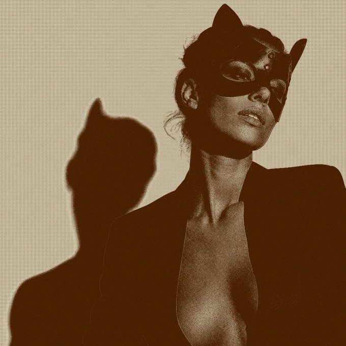 Sepia-toned portrait of a woman wearing a black cat-ear mask and an open blazer, with her shadow cast prominently against a plain wall behind her.