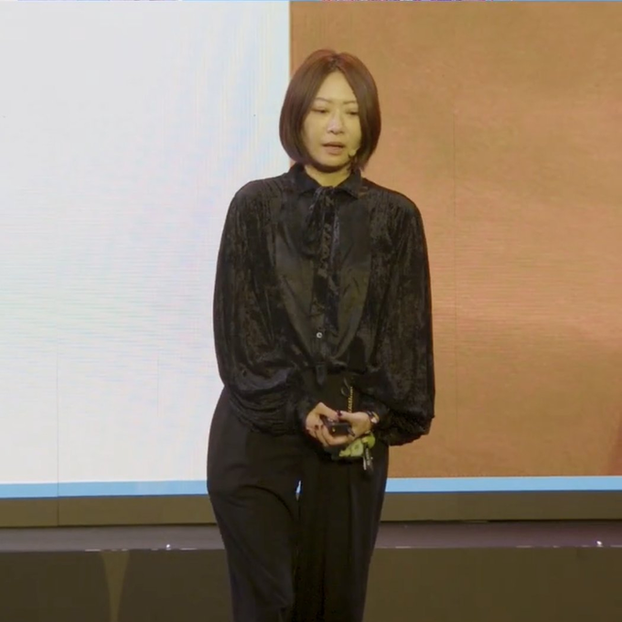 The image shows a woman standing on stage wearing a black blouse with a bow tie. She has short hair and is holding a cell phone in her hand, possibly taking notes or communicating with someone outside of the frame. The background features a large screen displaying text that reads "BREAKING: Hsiao-Wei Wang has officially stepped down as Co-Executive Director and Board Member of the Ethereum Foundation, effective today". This suggests that there might be an announcement or event related to the foundation's leadership change taking place on stage.
