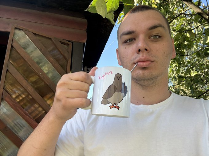 A man in a white t-shirt drinks from a mug labeled 'Курлык' featuring a pigeon illustration, with green leaves and a wooden structure in the background.