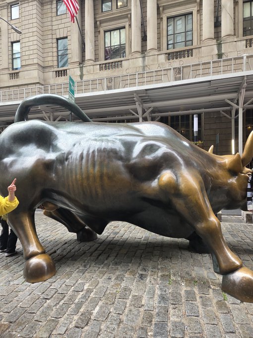 The Charging Bull bronze sculpture stands on a cobblestone street in front of a stone building, with a person in a yellow jacket gesturing toward it.