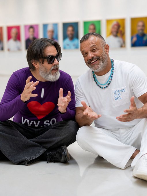Two men sit cross-legged on the floor in front of a colorful portrait series; one wears a purple "I ❤️ VVV.SO" shirt while the other sports a white "LOEWE L" tee with a turquoise beaded necklace. Their animated gestures and shared laughter echo the caption's "smile holds the supply doesn't," suggesting a celebratory moment amid a scarcity narrative tied to the VVV token.