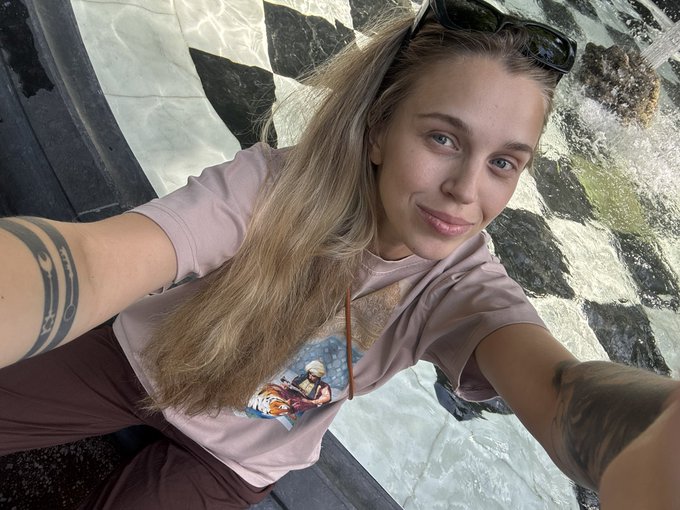 A selfie of a smiling young woman with blonde hair, sunglasses on her head, and a tattoo on her arm, wearing a pink t-shirt with an anime graphic.