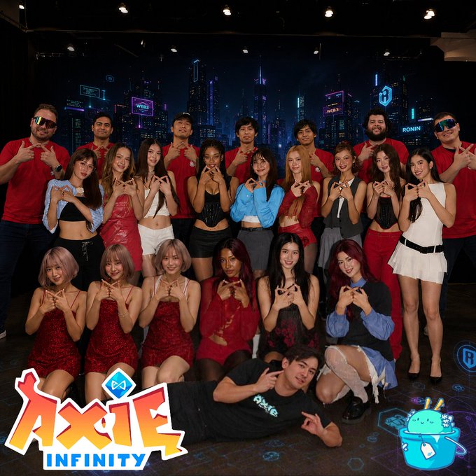Group photo of a large team posing with heart-shaped hand gestures on a stage with a cyberpunk city backdrop, featuring the 'Axie Infinity' logo in the bottom left and a cartoon Axie mascot in the bottom right.