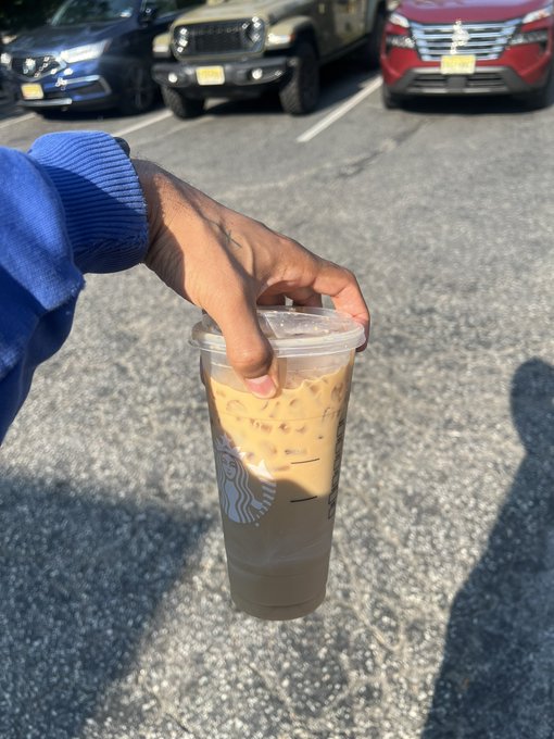 A hand holds a Starbucks iced coffee drink in a clear plastic cup, taken in a parking lot with several cars visible in the background.