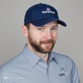 A man with a beard wearing a dark blue cap and a light blue button-up shirt poses for a portrait against a plain background.