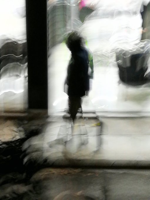 A blurred, impressionistic view through what appears to be wet glass shows a dark figure standing on a paved surface, with streaks of light and reflections distorting the scene.