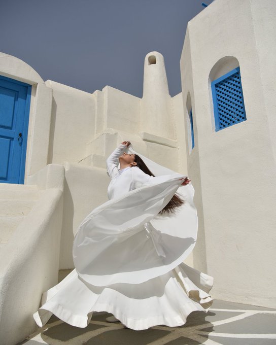 A woman in a flowing white dress poses dramatically among whitewashed walls, arches, and vivid blue doors and window grilles.