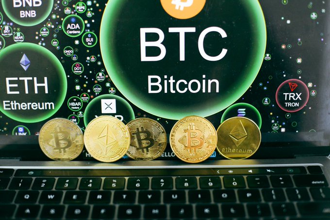 Close-up of physical cryptocurrency coins sitting on a laptop keyboard, with a screen displaying logos and tickers for BTC, ETH, BNB, ADA, TRX, DOT, and HBAR.