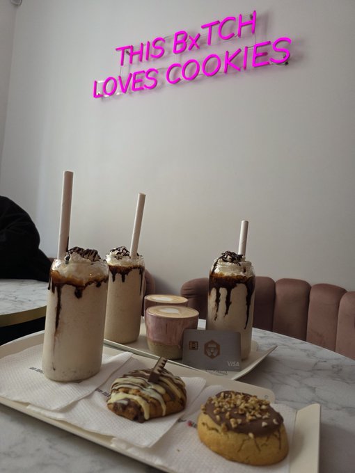 Three tall iced beverages with chocolate drizzle and two cookies sit on a marble table in a cafe, with pink neon text on the wall behind reading 'THIS B*TCH LOVES COOKIES'.