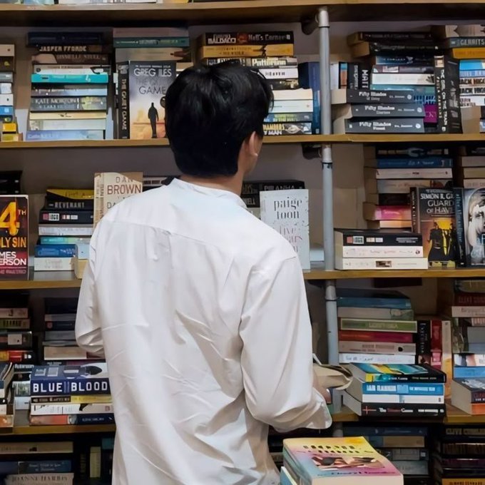 A person in a white shirt browses through densely packed bookshelves filled with novels by authors including Greg Iles, Dan Brown, and James Patterson.