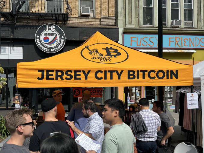 A crowd gathers under a bright orange "Jersey City Bitcoin" tent in front of an "F-C Academy Taekwondo Kickboxing" and "Fuss Friends" storefront, engaging with vendors at what appears to be an outdoor Bitcoin meetup or educational event. The image aligns with the post's description of community members learning about Bitcoin, capturing the lively street-level interest in cryptocurrency in Jersey City.