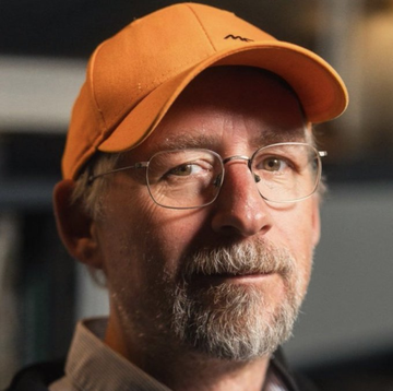A man wearing an orange baseball cap and wire-rimmed glasses, identifiable as cypherpunk and Blockstream CEO Adam Back, is shown in a close-up portrait. The image relates to the meme about his alleged standing buy order for all 21 million Bitcoin at $0.02 each, positioning him as a humorous "floor" for BTC's price.