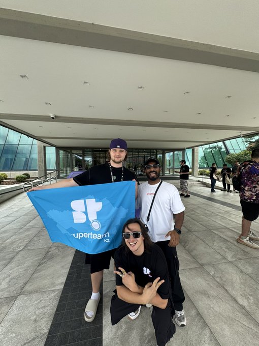 Three people pose outdoors at a modern building, with one person holding a blue flag reading 'Superteam KZ' featuring a stylized 'ST' logo.