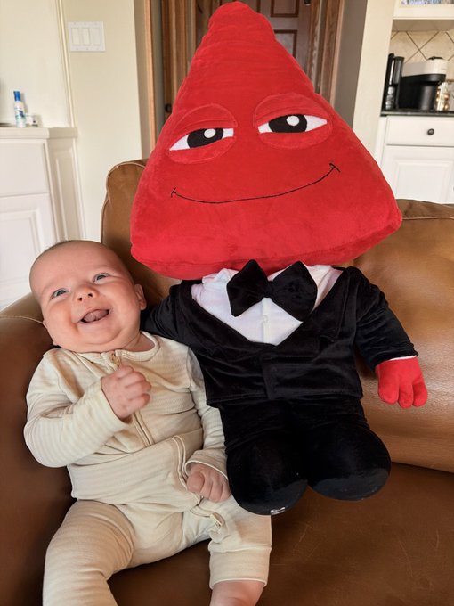 A smiling baby sitting on a leather couch next to a large plush mascot with a red triangular head, black suit, and bow tie.
