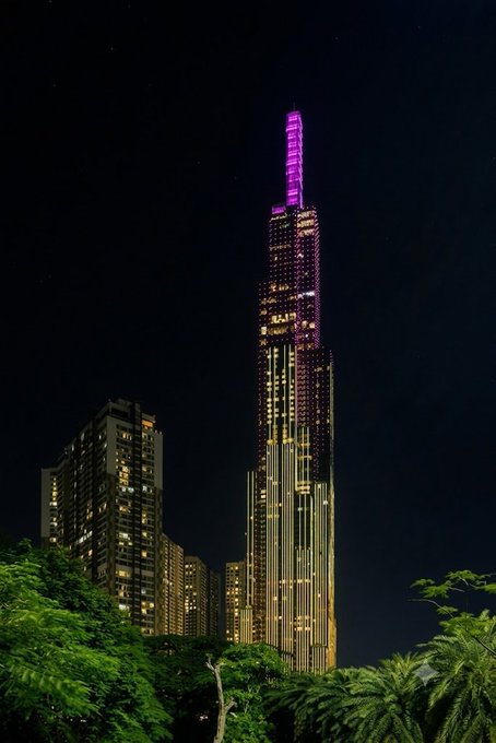 A nighttime photograph of the Landmark 81 skyscraper illuminated in purple and gold lights against a dark sky, framed by trees in the foreground.