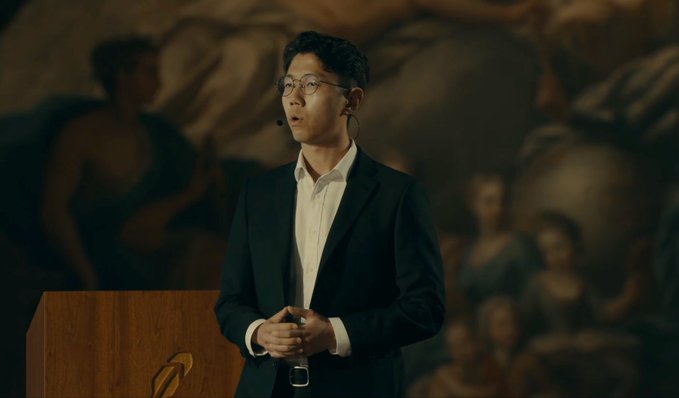 A young man in a dark suit and glasses speaks at a podium with a headset microphone, set against a dimly lit backdrop featuring classical-style artwork and an audience. The image accompanies a post about Robinhood ($HOOD) reaching over 1 million funded customer accounts outside the U.S., though no identifying text or logos are visible in the image itself.