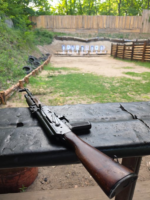 The image shows a gun resting on top of a wooden bench, with a backdrop of trees and a fence visible behind it. The gun appears to be an AK-47 or similar type of rifle, suggesting that this might be a shooting range or a training facility for marksmanship. The photo also includes the text "Thanks to everyone who came to the @MoneroKon shooting range side-event!", which implies that there was some sort of event or gathering related to the gun and its use at the location.