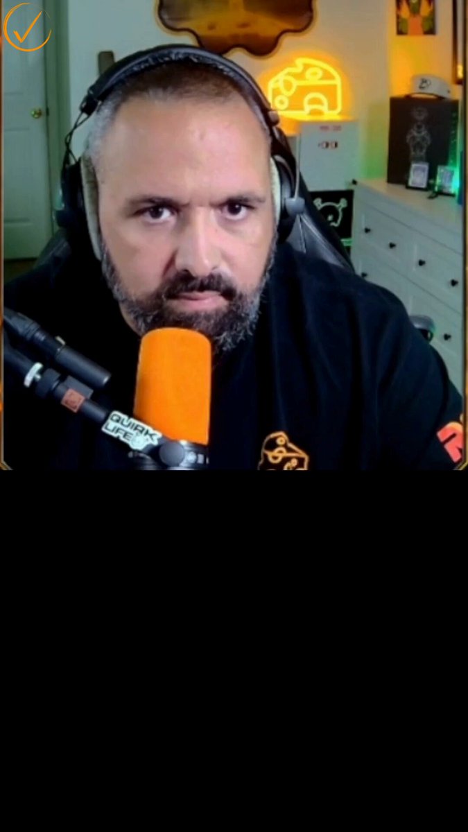 The image features a man wearing headphones sitting behind a microphone while recording a podcast. The man has a beard, and he appears to be focused on his work. In the background, there are two TVs mounted on the wall above the microphones. One of these TVs is turned off or in sleep mode.