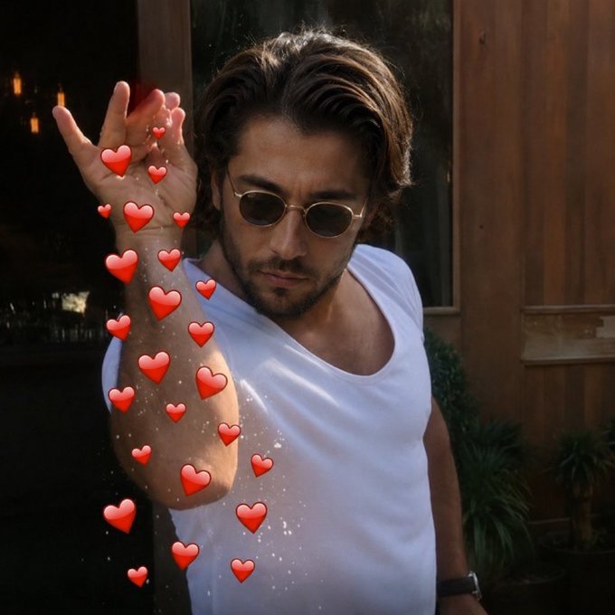 A man wearing sunglasses and a white tank top stands with his hand raised, surrounded by floating red heart emojis.
