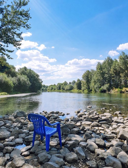 A bright blue plastic chair sits on a rocky riverbank overlooking calm water and dense green trees beneath a partly cloudy blue sky. The serene outdoor scene does not appear directly related to the accompanying post about Base Batches Week 3, which describes founder and product-building work and discussions with the Base ecosystem team.
