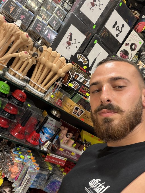 A bearded man takes a selfie inside an adult novelty store surrounded by shelves of packaged merchandise, with a "call girl" DVD visible among the products, visually capturing the "memecoin supercycle" hype and the chaotic, anything-goes energy of the crypto market pump.