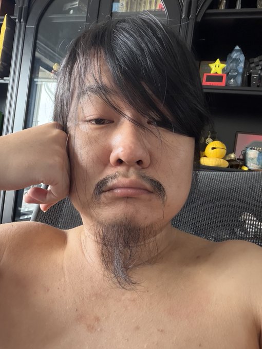 A shirtless man with long dark hair covering part of his face and a goatee rests his head on his hand, posing for a selfie in a room with shelves displaying various items.