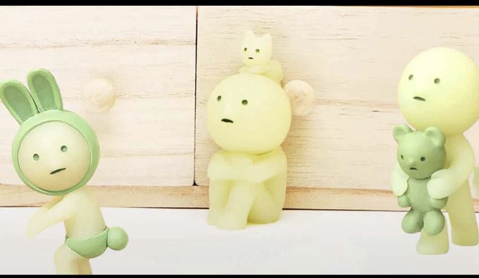 Three small, pastel-green figurine characters with blank, slightly worried expressions are arranged together against a light wooden backdrop, with the central figure hugging a tiny bear and another perched on its head. The whimsical, soft-toned scene pairs with the post's friendly greeting and Bitcoin address, giving a gentle, community-oriented feel to the message.