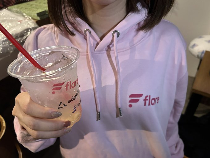 A person wearing a light purple "flare" branded hoodie holds a clear plastic cup displaying the "Flare" logo along with partners like edodev and PUR. The image showcases branded merchandise, matching the post about the abundance of Flare merch available.
