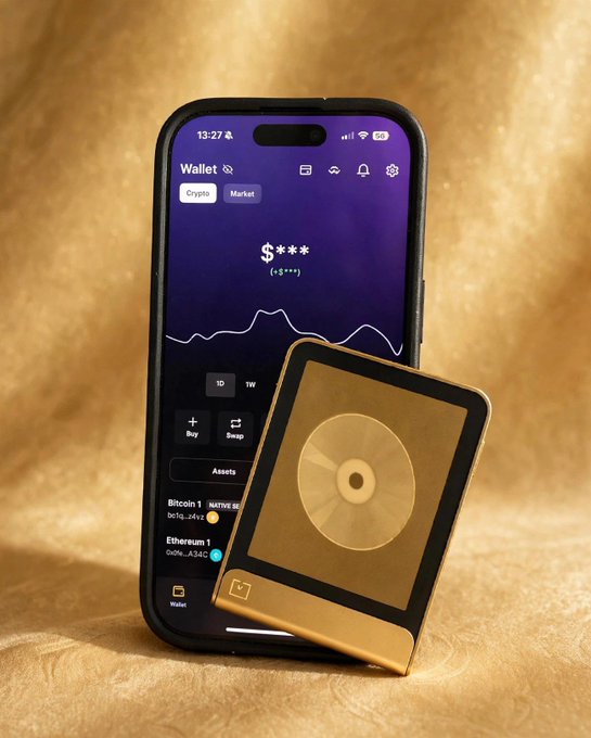 A smartphone displaying a crypto wallet app with a chart, 'Buy' and 'Swap' buttons, and listings for 'Bitcoin 1' and 'Ethereum 1', propped up next to a gold hardware wallet device on a textured gold surface.