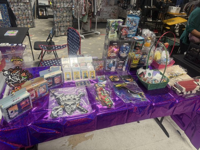 A vendor's table draped in a shiny purple tablecloth, displaying various anime and pop-culture merchandise including trading cards, t-shirts, toys, and a gift basket.