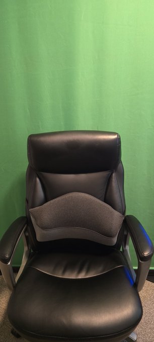 A black leather office chair with a lumbar pillow sits in front of a green screen backdrop.