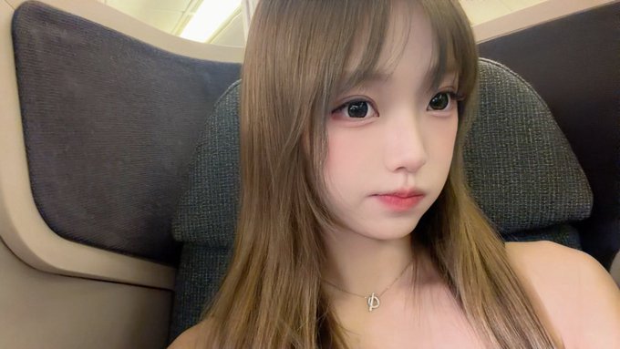 A selfie of a young woman with long brown hair wearing a pink top and a necklace, seated in what appears to be a vehicle or airplane cabin.