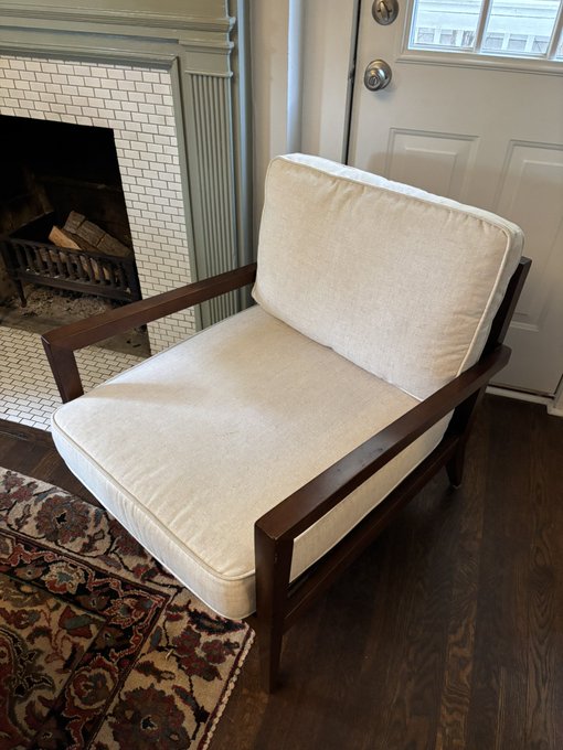 A cream-colored cushioned armchair with a wooden frame positioned next to a white tiled fireplace and a paneled white door, sitting on dark hardwood flooring with a patterned rug.