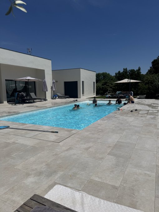 A group of people enjoys a sunny day swimming in a rectangular pool beside a modern white house, with umbrellas providing shade around the patio. The scene matches the post's invitation to "touch the water" with its clear blue pool and relaxed summer vibe.