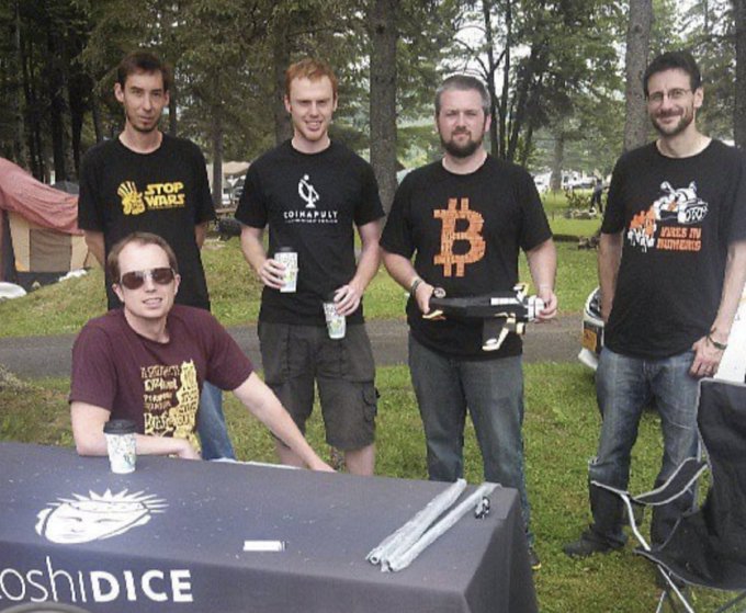 A group of five men pose at an outdoor table with a Satoshidice banner, wearing t-shirts featuring logos like Stop Wars, Coinapult, Bitcoin, and Bitcoin Not Bombs.