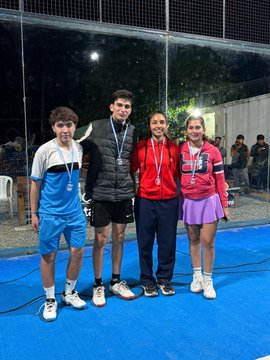 The image features four people standing together on a blue surface, posing for a photo. They are all wearing medals around their necks, indicating that they have won an event or competition. The group consists of two men and two women, each holding a medal in front of them. In the background, there is a speaker system visible, suggesting that this might be a sports event or a similar gathering where music was played to entertain the attendees.
